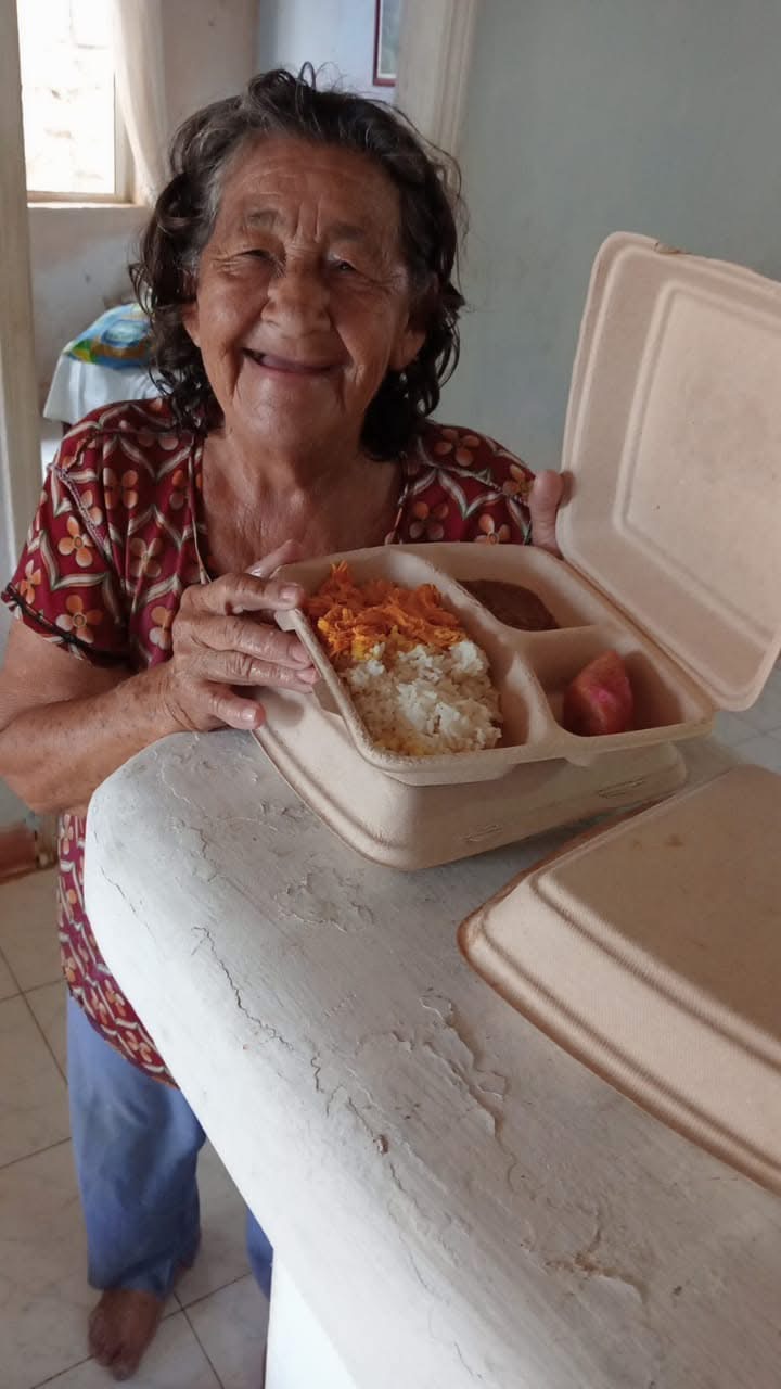 Mujer mayor sonriendo mientras recibe una porción de comida en un recipiente biodegradable.